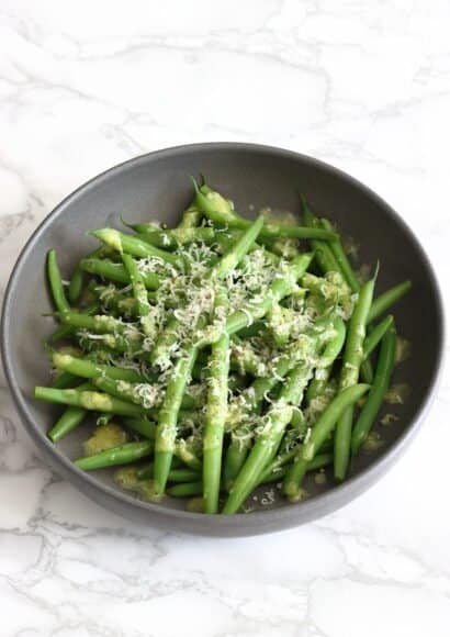 Warm green beans with lemon and parmesan dressing served in a dark grey bowl on a marble worktop, finished with finely grated parmesan, sea salt and black pepper.