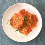 A bowl of 4 golden tomato fritters flecked with feta cheese and sprinkled with chopped mint sitting on a bed of yoghurt and mint. The bowl is sat on a moody blue textured background board.