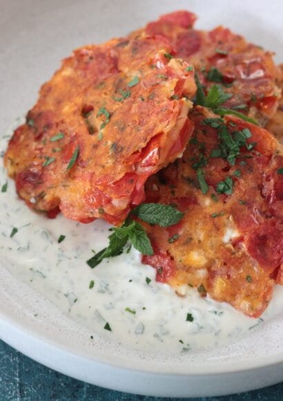 A close up of a bowl of 4 golden tomato fritters flecked with feta cheese and sprinkled with chopped mint sitting on a bed of yoghurt and mint