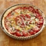Overhead square crop of a golden tomato tart with mustard shallots, alternating red and yellow tomato slices arranged in circles, topped with melted cheese and scattered thyme. The tart is still in its fluted tin, and is sat on a chopping board just out of the oven.