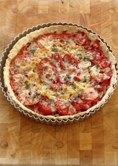 A metal fluted tart tin, sat on a wooden board, containing a cooked tomato tart tart with mustard shallots. The tomatoes are layered in circles with red and yellow tomatoes, topped with golden melted cheese and flecked with thyme leaves.