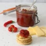 A stack of crackers in the foreground with a wedge of cheddar on the top one which has a big spoon of tomato and chilli jam on the top. This is sat in front of a small pile of sliced cheddar, a large pot of the jam with a spoon in it, and a handful of cherry tomatoes and a red chilli.