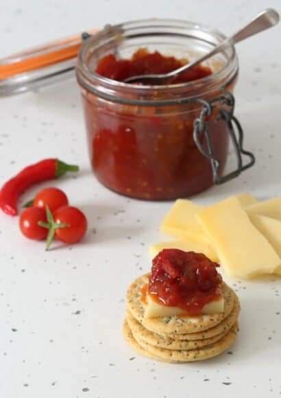 A stack of crackers in the foreground with a wedge of cheddar on the top one which has a big spoon of tomato and chilli jam on the top. This is sat in front of a small pile of sliced cheddar, a large pot of the jam with a spoon in it, and a handful of cherry tomatoes and a red chilli.