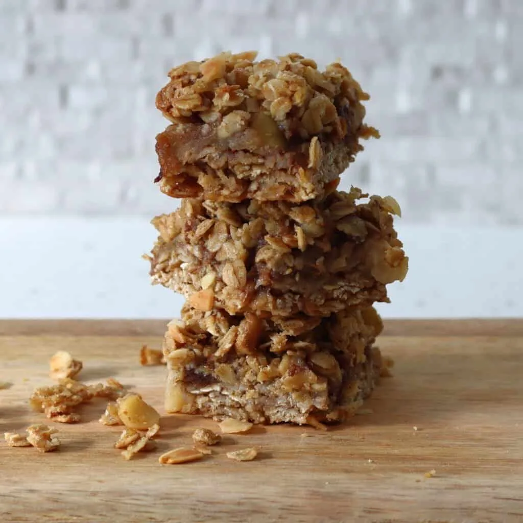 A stack of 3 apple nut and crumble bars cut into squares, placed on a board, with a scattering of oats and chopped nuts around
