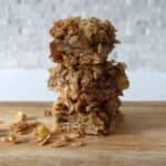 A stack of 3 apple nut and crumble bars cut into squares, placed on a board, with a scattering of oats and chopped nuts around