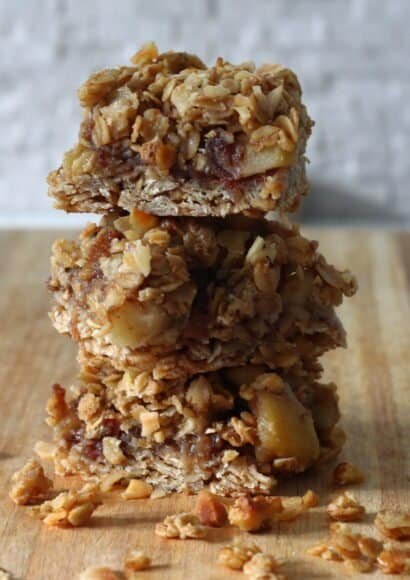 A stack of 3 apple nut and crumble bars cut into squares, placed on a board, with a scattering of oats and chopped nuts around