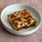 A bowl containing baked moussaka with golden béchamel topping showing the layers of lamb, lentils, vegetables and aubergine.