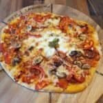 Homemade pizza on a wooden circular board, topped with mushrooms, olives, peppers, melted mozzarrella, and the important pizza sauce spread on the dough.