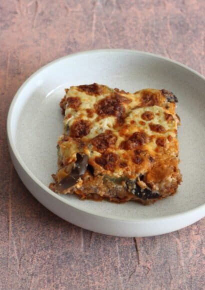 A bowl containing baked moussaka with golden béchamel topping showing the layers of lamb, lentils, vegetables and aubergine.