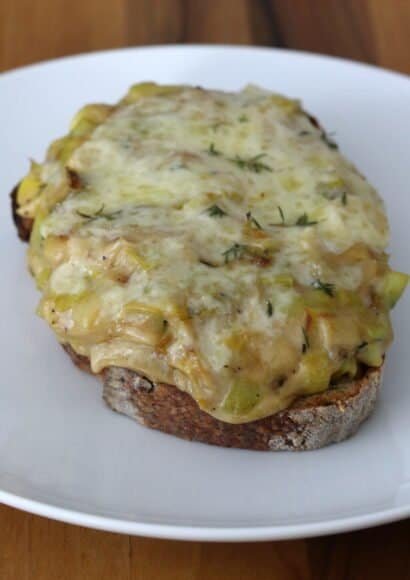 A slice of cheesy leek rarebit on toast with melted sauce oozing over the edge sprinkled with fresh thyme leaves.